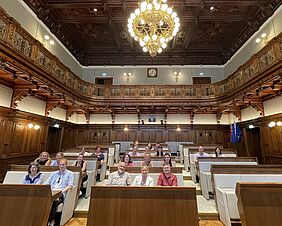 Zu sehen sind sieben Menschen in einem Halbkreis um einen schwarzen Konferenztisch. Die Gruppe wirkt konzentriert, einige hören zu, eine Person in der Mitte spricht gestikulierend. An der Wand hinter ihnen hängen viele gerahmte Fotos und ein großer Bildschirm. Die Personen befinden sich in einer Besprechung oder einer Diskussionsrunde. 