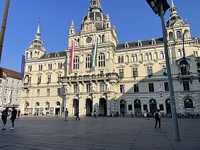Zu erkennen ist ein großes historisches Gebäude mit reich verzierter Fassade und mehreren Türmen. Es handelt sich um das Rathaus von Graz, erkennbar an den Flaggen – unter anderem der österreichischen, der EU-Flagge und einer grün-weißen Landesfahne. Vor dem Gebäude befinden sich einige Menschen. An der Fassade hängen Blumenkästen, im Erdgeschoss sind Läden und eine Bankfiliale zu sehen. 