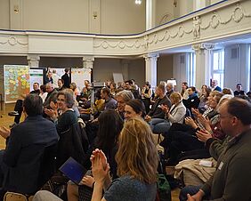 Zu sehen ist ein opulenter Saal, ausgestattet mit hintereinander aufgereihten Stühlen, die alle besetzt sind. Es wirkt voll wie in einem Theatersaal zur Premiere eines Stücks. Im Hintergrund sind Tafeln und Flipcharts mit Plakaten und bunten Notizzetteln zu erkennen. Das Publikum applaudiert.