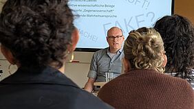 Ein Mann sitzt vor einer Klasse, deren Köpfe im Bild von hinten zu erkennen sind. Der Mann trägt eine Brille und Glatze. Hinter ihm befindet sich eine Leinwand, die eine Präsentation abzeichnet.