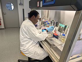 Zu sehen ist ein Wissenschaftler in einem weißen Kittel, der in einem sterilen, grauen Labor arbeitet. Vor ihm liegen verschiedene Laborutensilien. Der Mann trägt blaue Gummihandschuhe. 