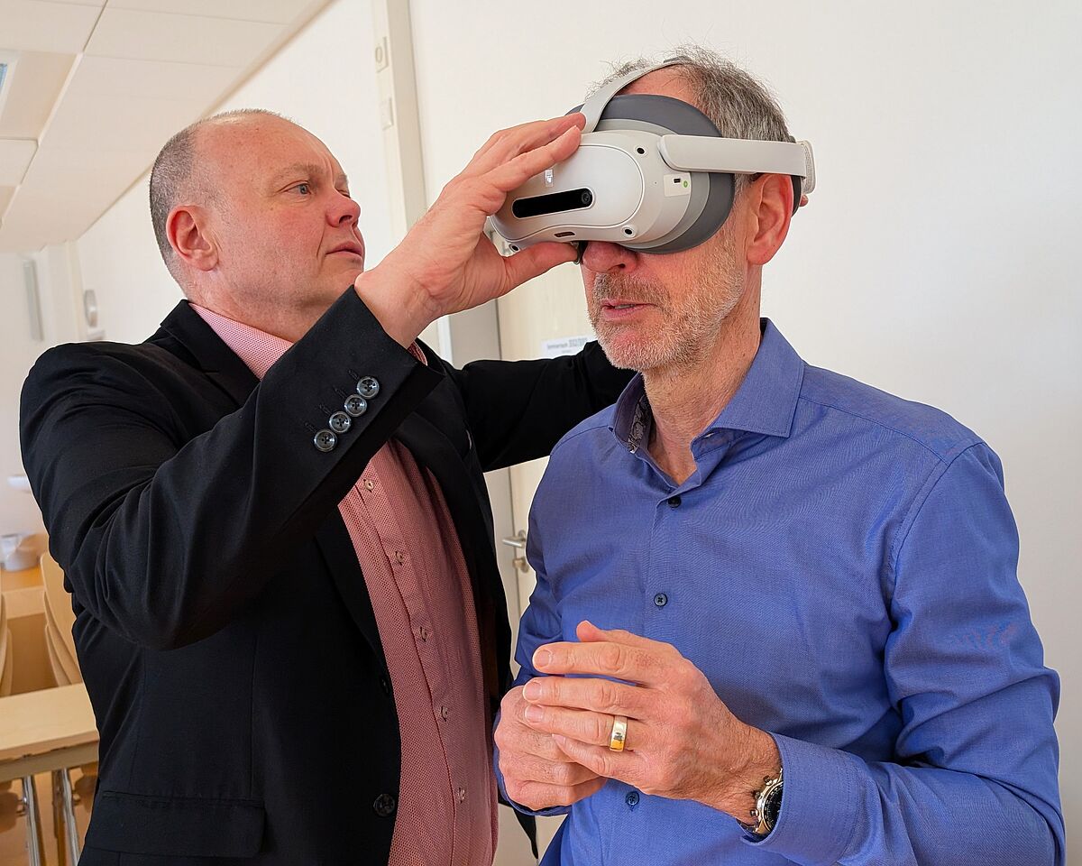 Ein Mann in einem schwarzen Sakko legt einem anderen Mann eine weiße VR-Brille an.