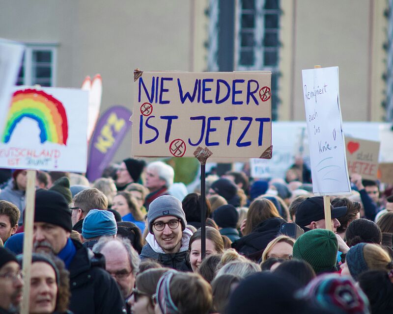Auf dem Bild sind demonstrierende Menschen zu sehen. Auf dem Schild einer Person steht "Nie wieder ist jetzt".